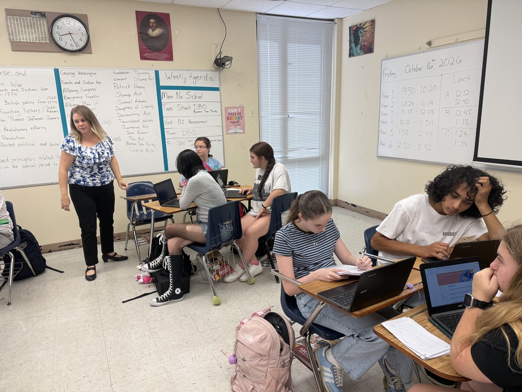 Students seated in classroom chairs, working on laptops; one student stands at a whiteboard, others look at screens.