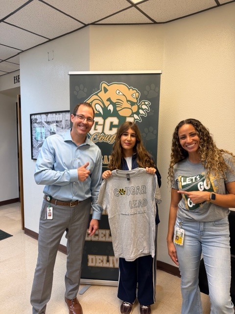 Three people stand in front of a banner with a school mascot. Two hold up a gray shirt.