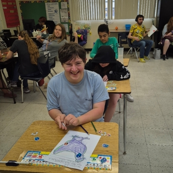 A classroom with students working at desks. A boy smiles at the camera, holding a marker.
