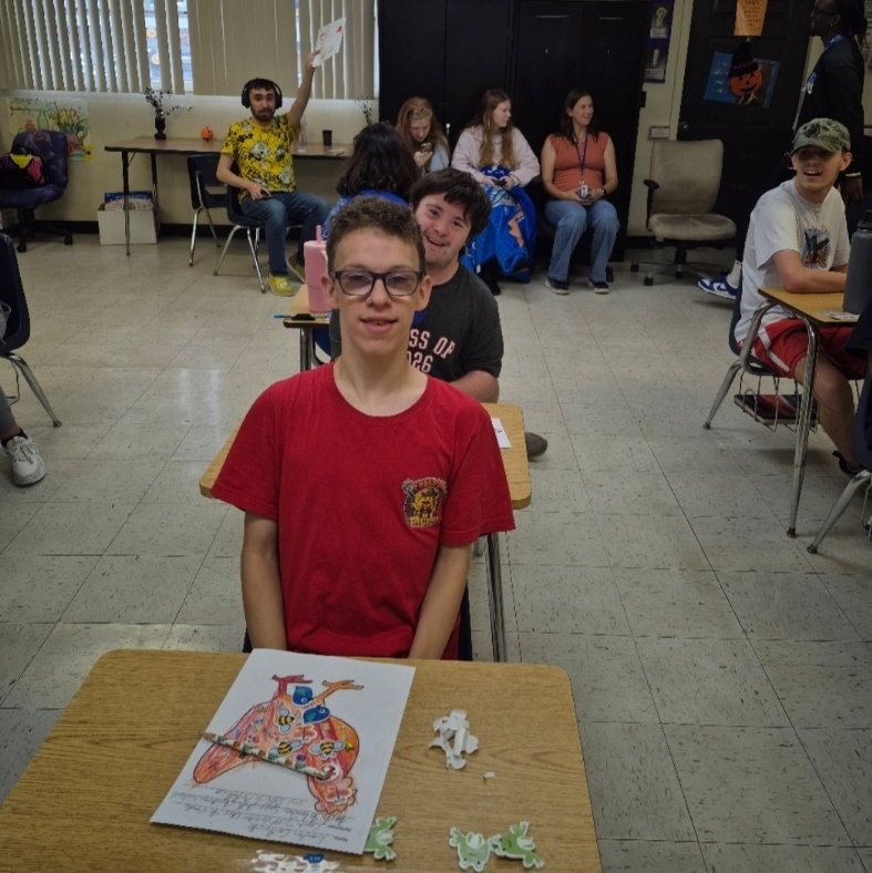 Several students seated at desks in a classroom. One student in red stands in front of his desk with artwork.