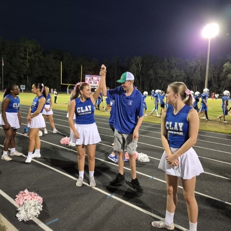 Adults and young people stand on a track field, with cheerleaders holding pom-poms, under bright lights at night.