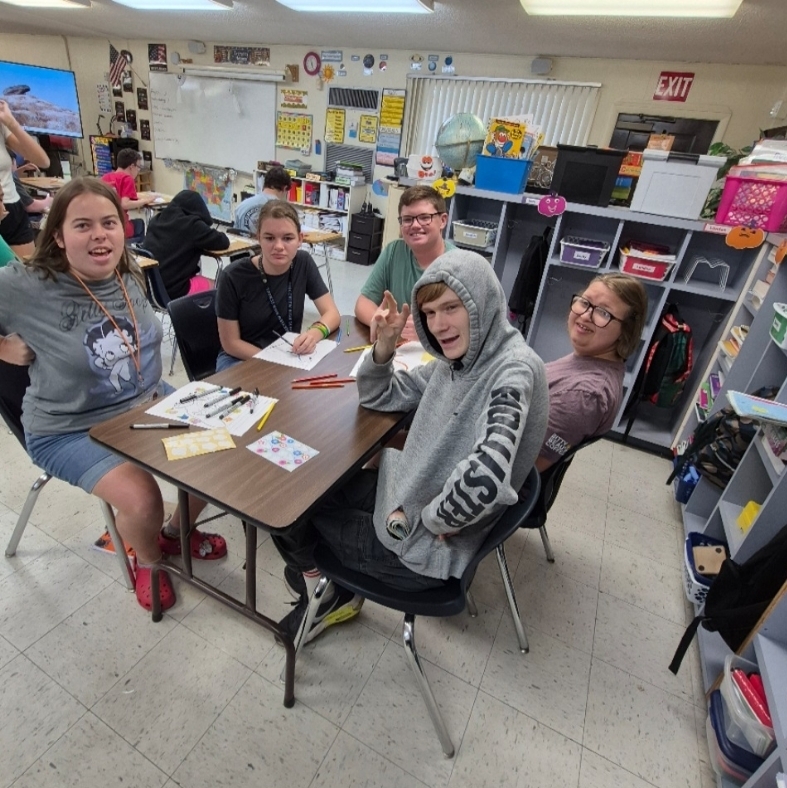 Five students seated around a table with papers and pens, smiling at the camera. A classroom background includes shelves with storage bins and a whiteboard.
