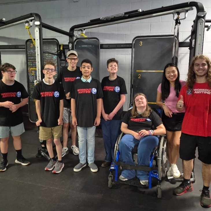 Nine students in sports gear in a gym, with one student in a wheelchair, smiling.