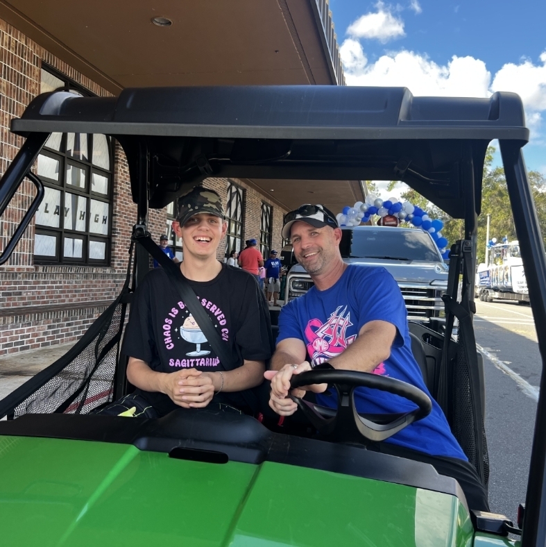 Two people in a golf cart outside a brick building. One person wearing a hat sits in the front.