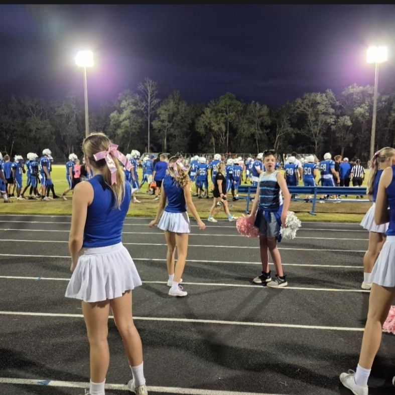 Cheerleaders in blue tops and white skirts stand on a football field. A man in a hat and sneakers stands near them.