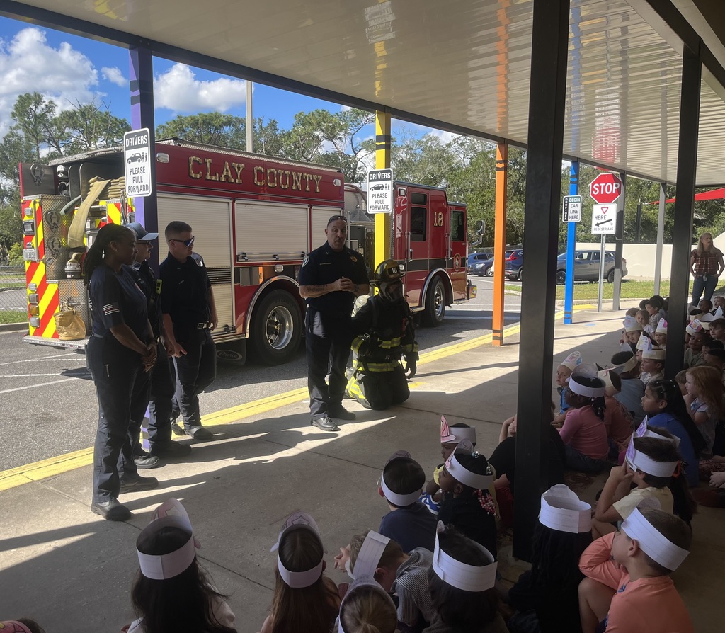People in uniform stand in front of a firetruck; seated people wear white headbands and watch.