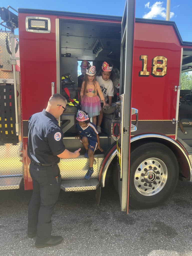 A fire truck is open. A firefighter helps kids into the truck. One child wears a hat.