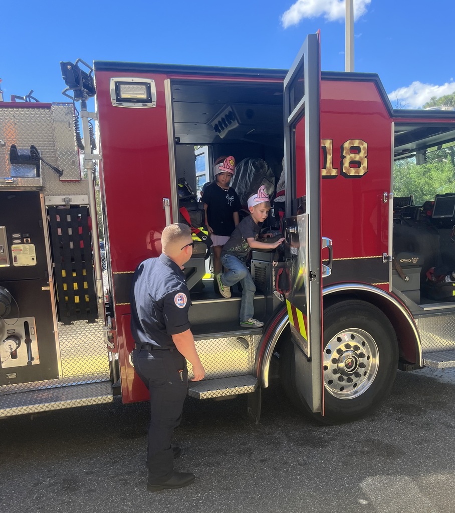 Man in uniform helps two children climb stairs of open fire truck, numbers 18 on door.