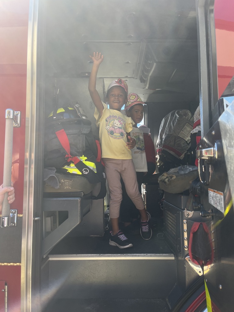 Two kids stand inside a firetruck with one child waving. A helmet rests on a shelf.