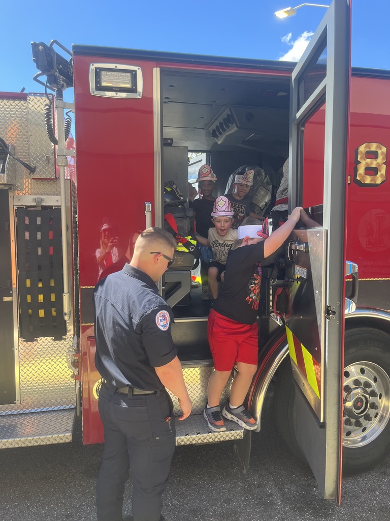 Police officer standing by fire truck, children inside, one climbing up ladder, another wearing hat.