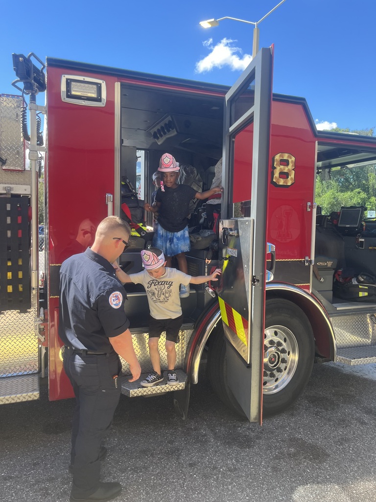 A firefighter helps two children climb into a fire truck with open doors on a sunny day.