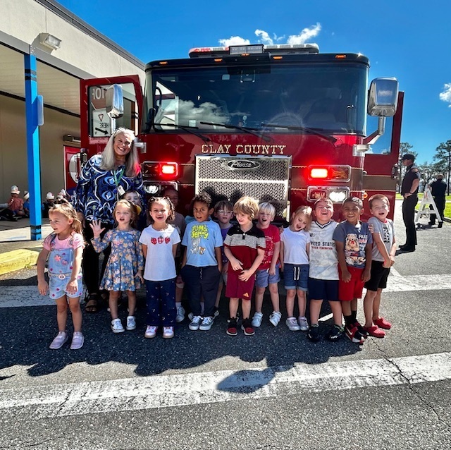 A group of children and an adult stand in front of a red firetruck. They are on a paved road.