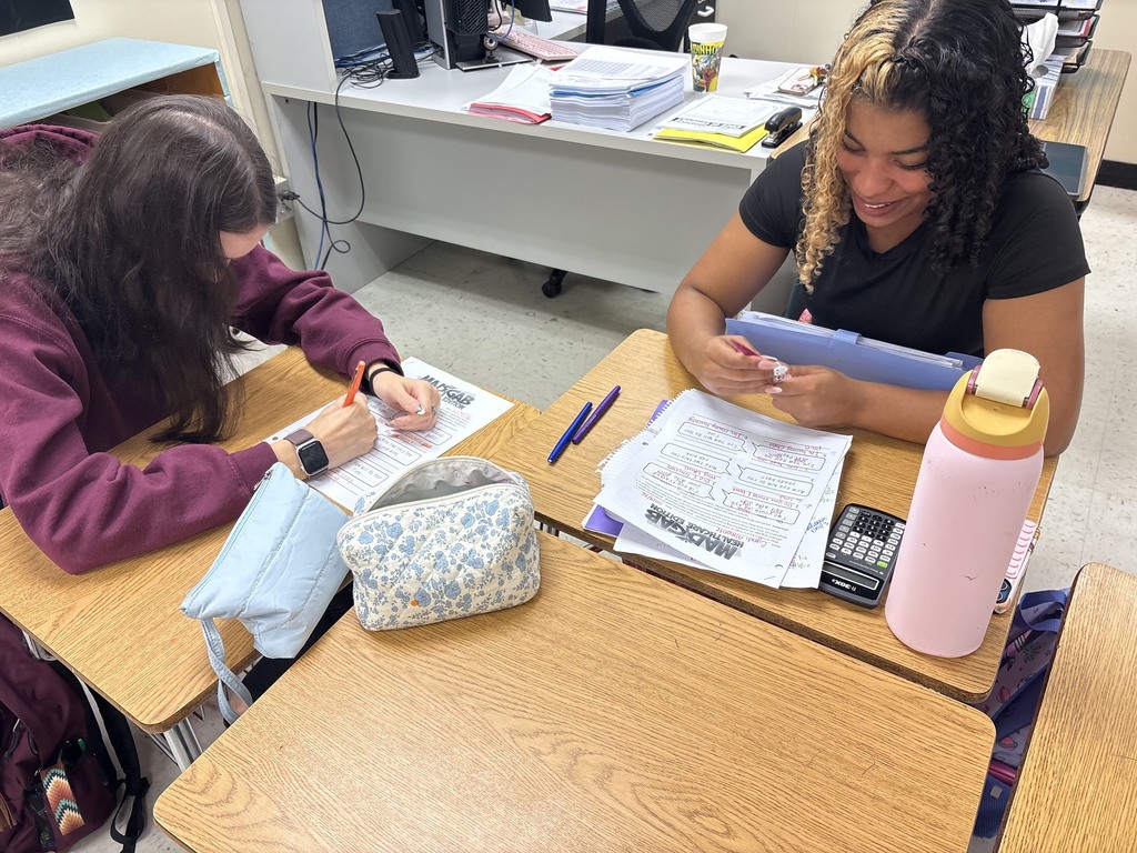 Two students seated at desks, each writing on paper with pens, with one using a calculator.