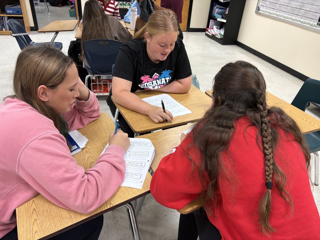 Three girls in a classroom are seated at desks, one writing while the others listen intently.