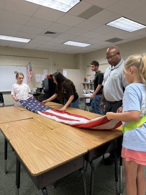 Several children help fold an American flag on a table while adults stand nearby.