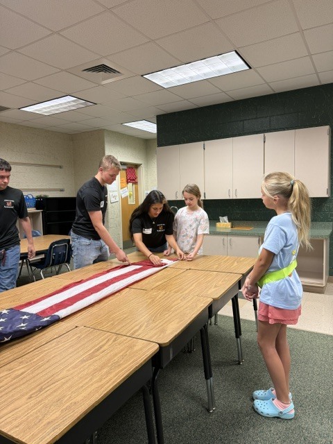 Group of people standing around tables, one holding a flag, others watching, in a classroom.