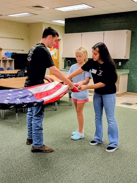 Two students helping a man carry a flag in a room with tables and a kitchen.