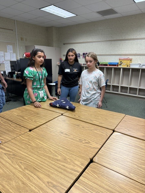 Three students stand around a large table with a folded flag on it inside a classroom.