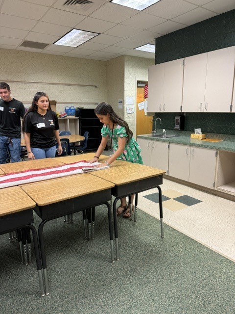 Three people in a classroom with desks and a counter. One person is kneeling over a desk.