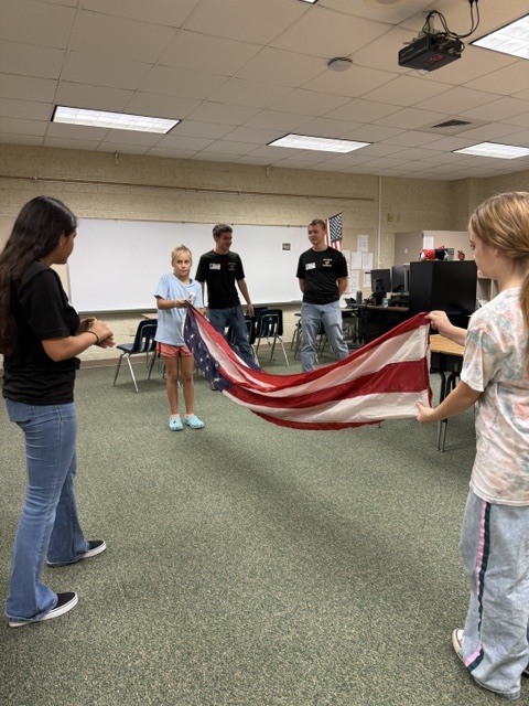 People unfold a large flag in a classroom with a projector on the ceiling.