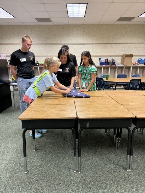Four people stand around a table folding a blue fabric. The table is surrounded by other tables and chairs.