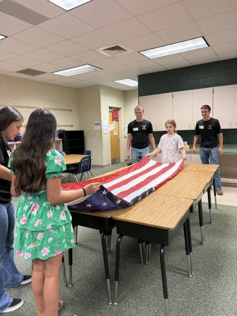 Five students stand around a table, holding an American flag. A woman in a floral dress folds the flag.