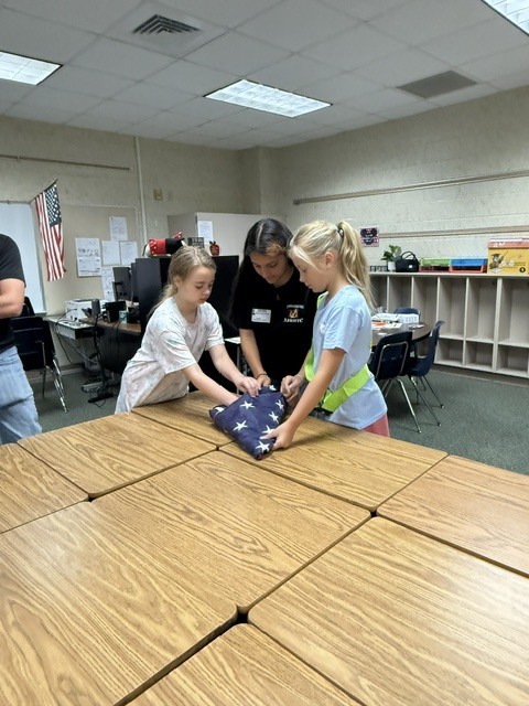 Two children folding an American flag on a table in a classroom. A teacher assists.