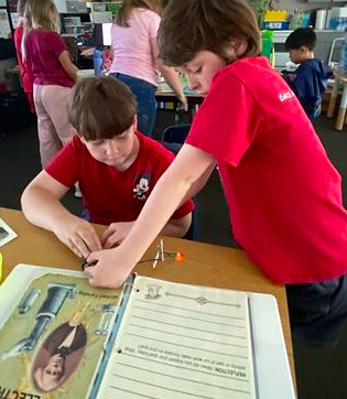Two boys working on a project in a classroom. One boy is seated, the other standing, both wearing red shirts.