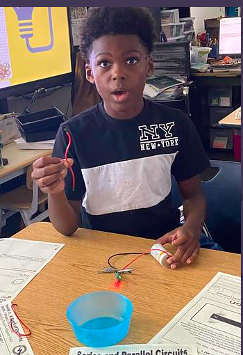 A young boy seated at a table holds wires and a battery, with blue liquid in a small bowl.