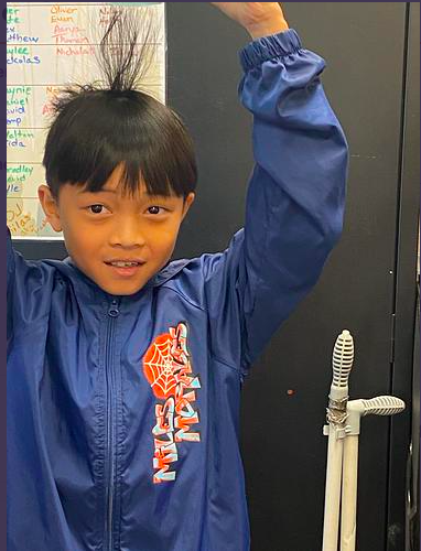 Young boy with spiky hair, wearing a blue raincoat with a spider design, raising one arm.