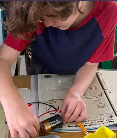 Student hands on a desk, holding a battery and pencil, working on a circuit with wires.