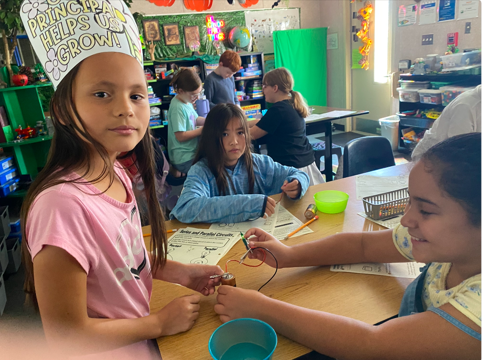 Students in a classroom, one wearing a paper chef hat, work on a project at a desk.