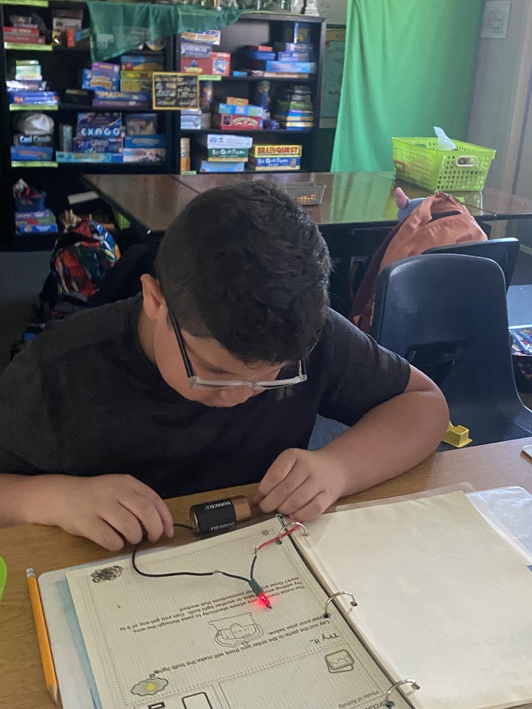 A student sits at a desk, focused on a project with wires and a light.