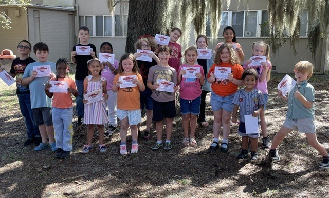 A group of children holding certificates stand outside, smiling in front of a tree and buildings.