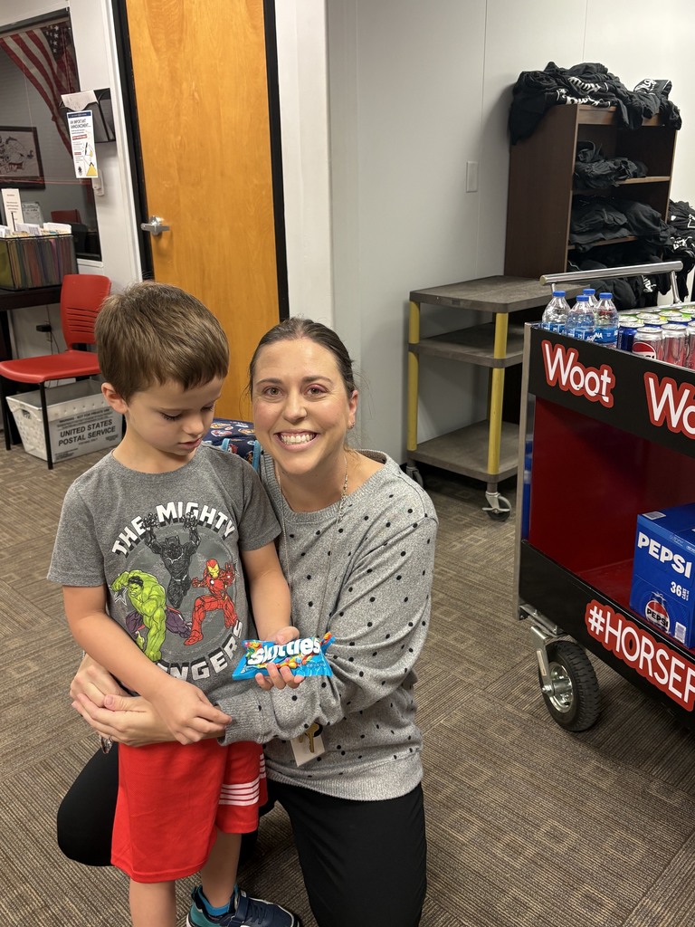 Woman kneeling holds boy wearing red shorts. Boy holds small toy. Red wheeled cart filled with items.