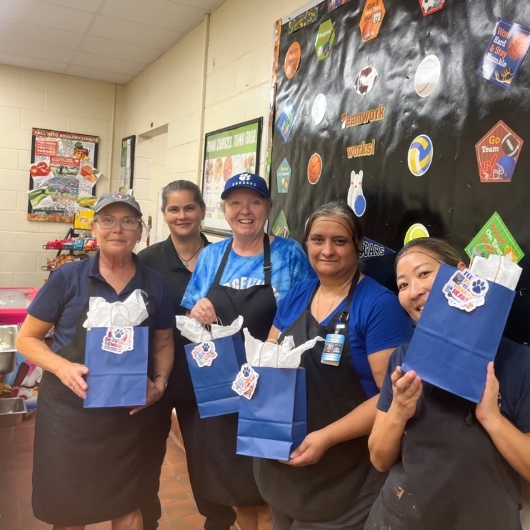Five women in aprons hold blue bags. One woman holds a blue bag with a white ribbon.