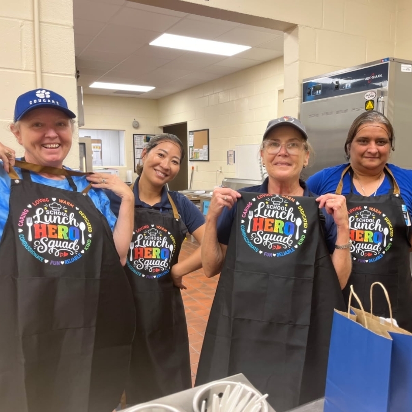Four people in aprons stand together in a cafeteria. Three wear hats and one wears glasses.
