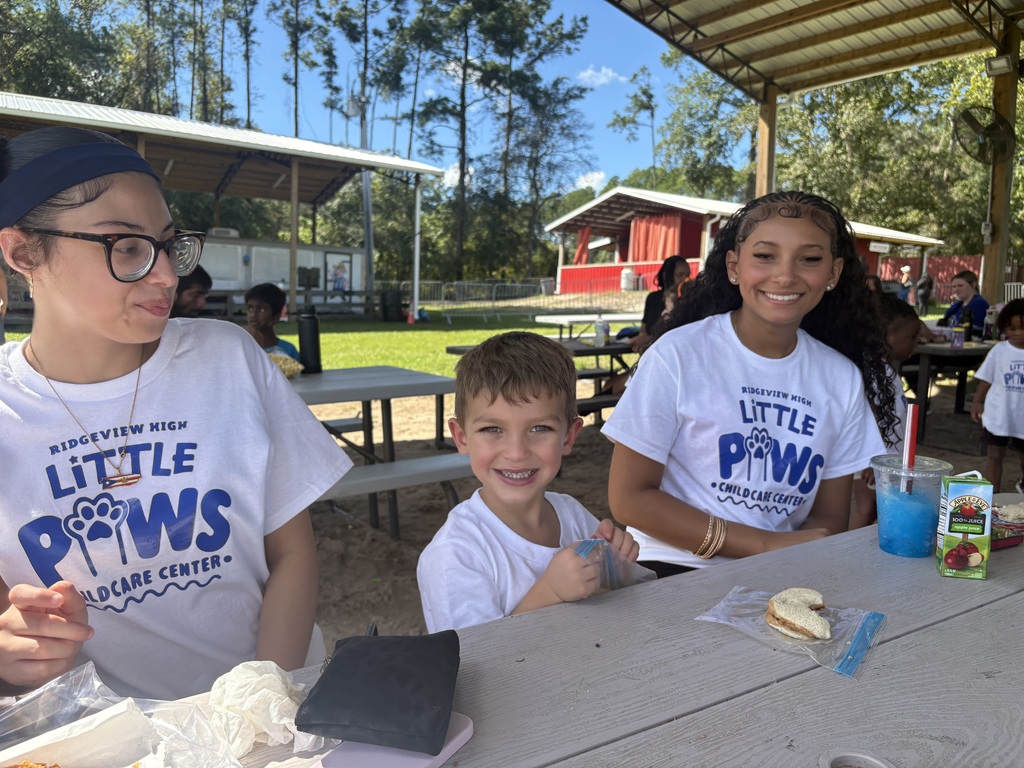 Three students at an outdoor picnic table, one holding a drink, smiling, and surrounded by food.