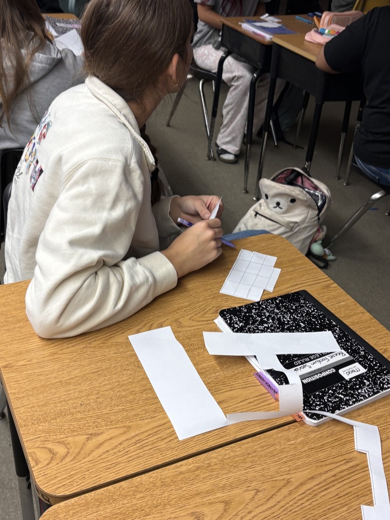 Students seated in a classroom, one cutting paper, and several desks with backpacks and notebooks.