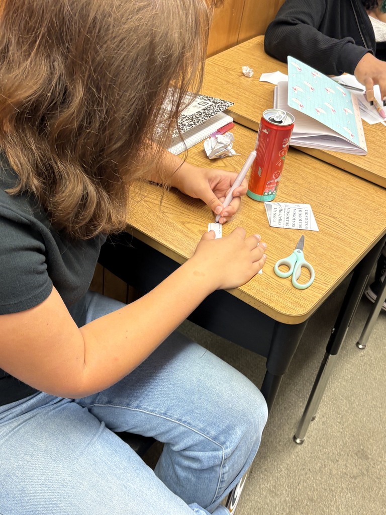 Student seated at a desk holds scissors and a pen while cutting paper, with a cup and book nearby.
