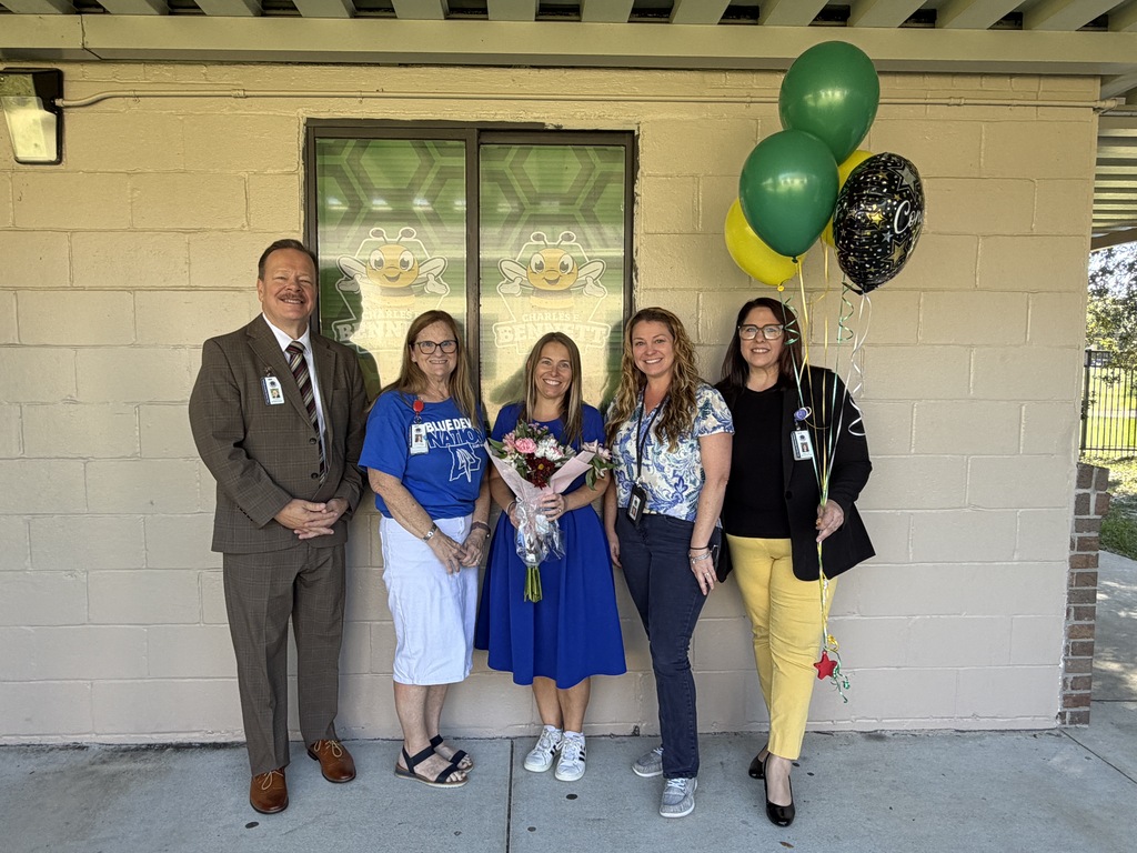 Five people stand in front of a building. Two hold balloons. Two have flowers. One wears a suit and tie.