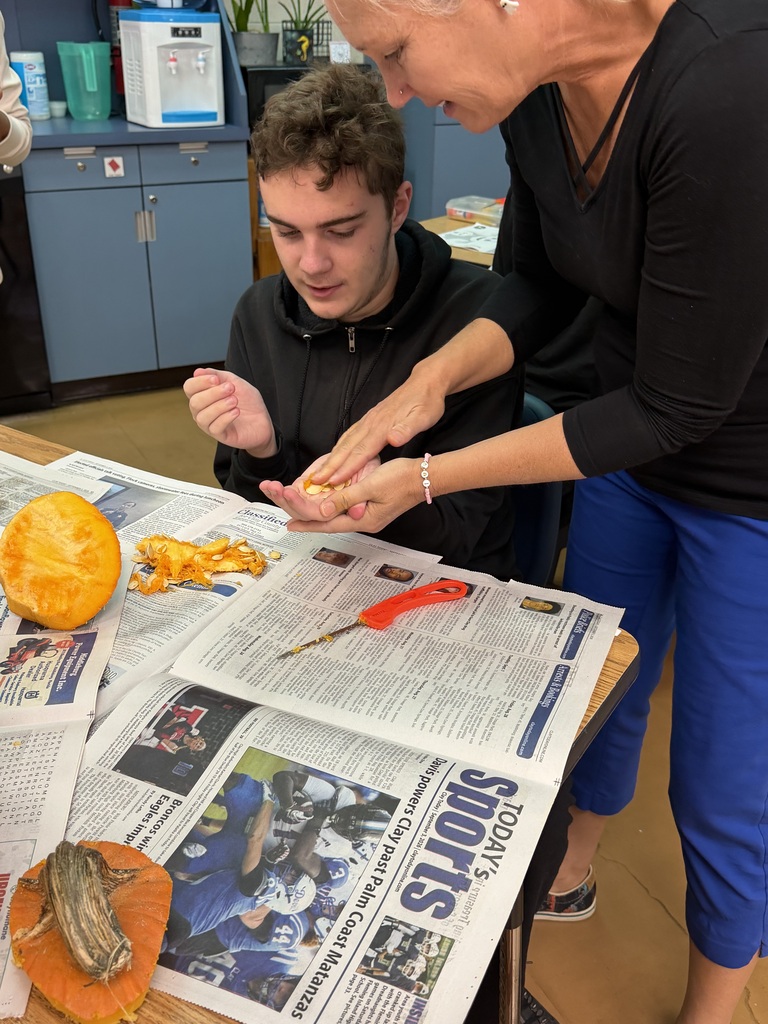 Woman helping student with carving a pumpkin on a table with newspaper. Various objects on table.