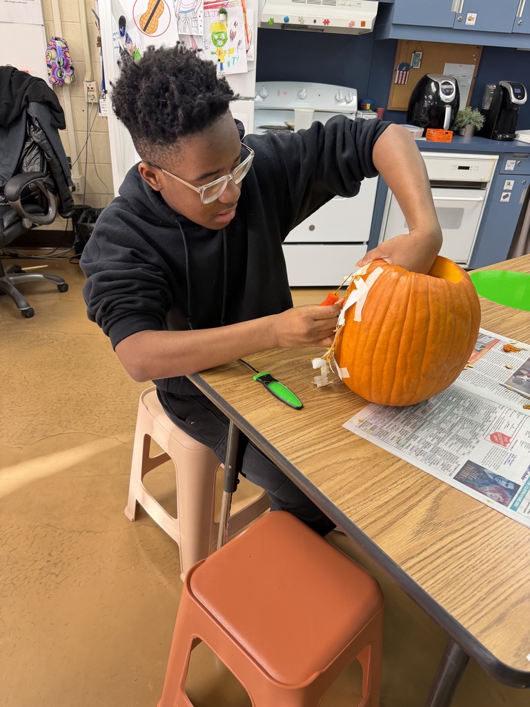A student wearing glasses and a black hoodie carves a pumpkin on a table.