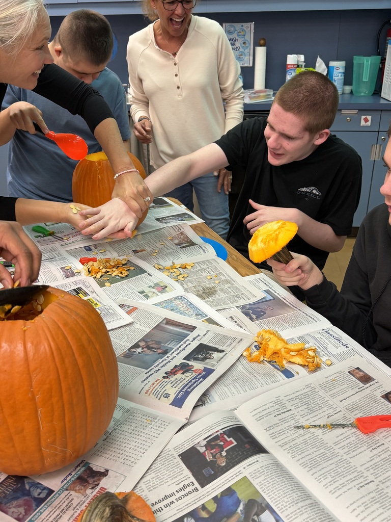 Students and teachers work together to carve pumpkins on a table covered in newspapers.