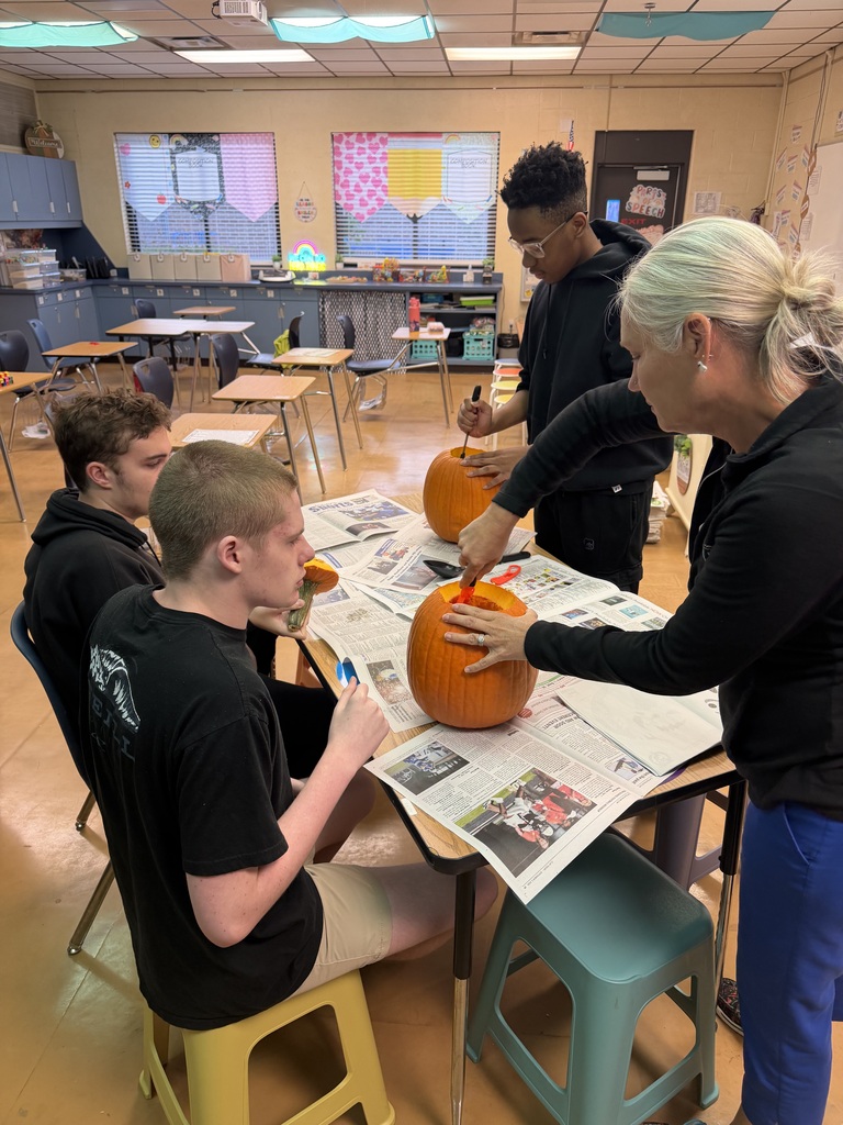 Four people in a classroom. Three are seated around a table with pumpkins. One is holding a knife.