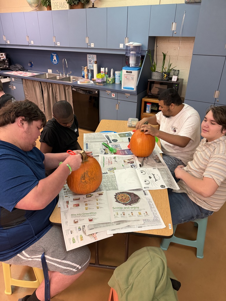 Four people sit around a table, carving pumpkins with tools. A sink and cabinets are in the background.