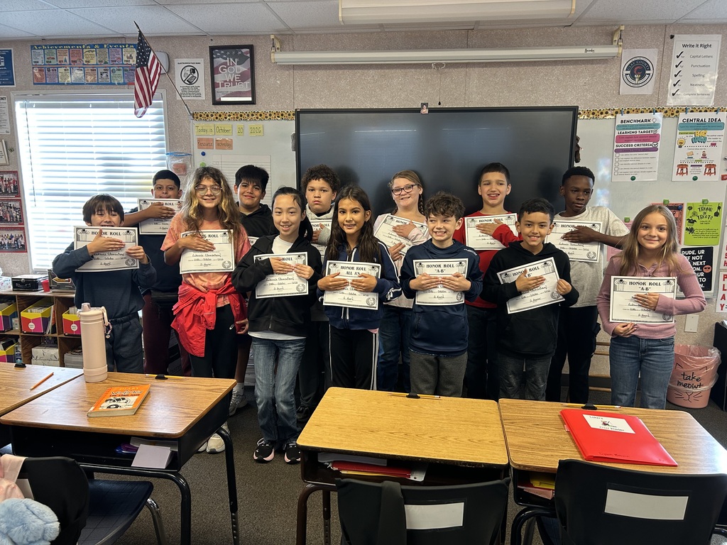 Children stand in a classroom with certificates, holding them up. Behind them are a whiteboard and a large screen.