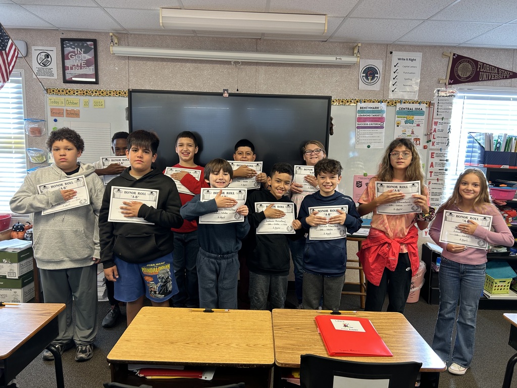 Group of students in a classroom holding certificates, standing around a table with chairs and a flag.