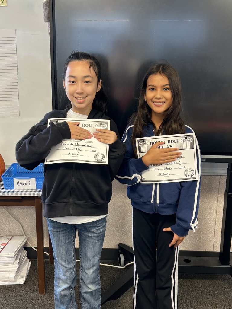Two young girls holding certificates with smiles in a classroom, a table with books behind them.