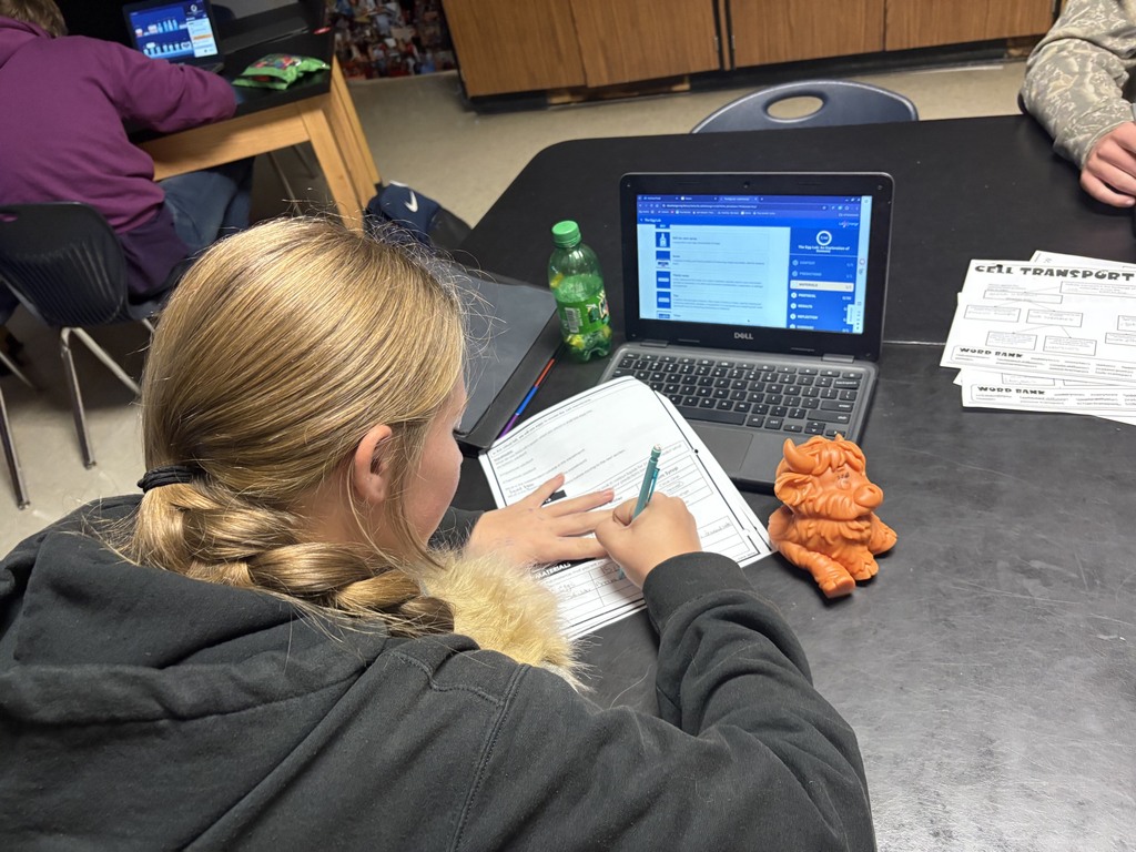 A student writes on paper at a table. A laptop and plastic bottle sit on the table.